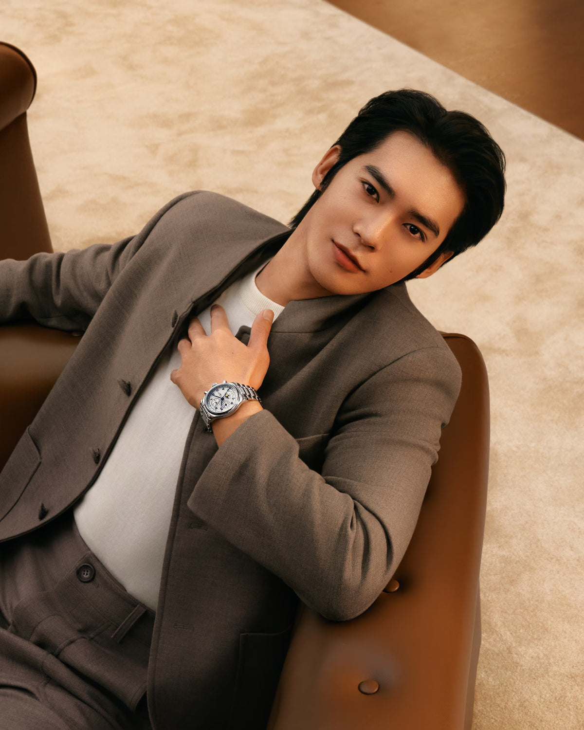 A man in a brown suit and white shirt reclining on a brown leather chair, wearing a silver-toned wristwatch with a white dial.