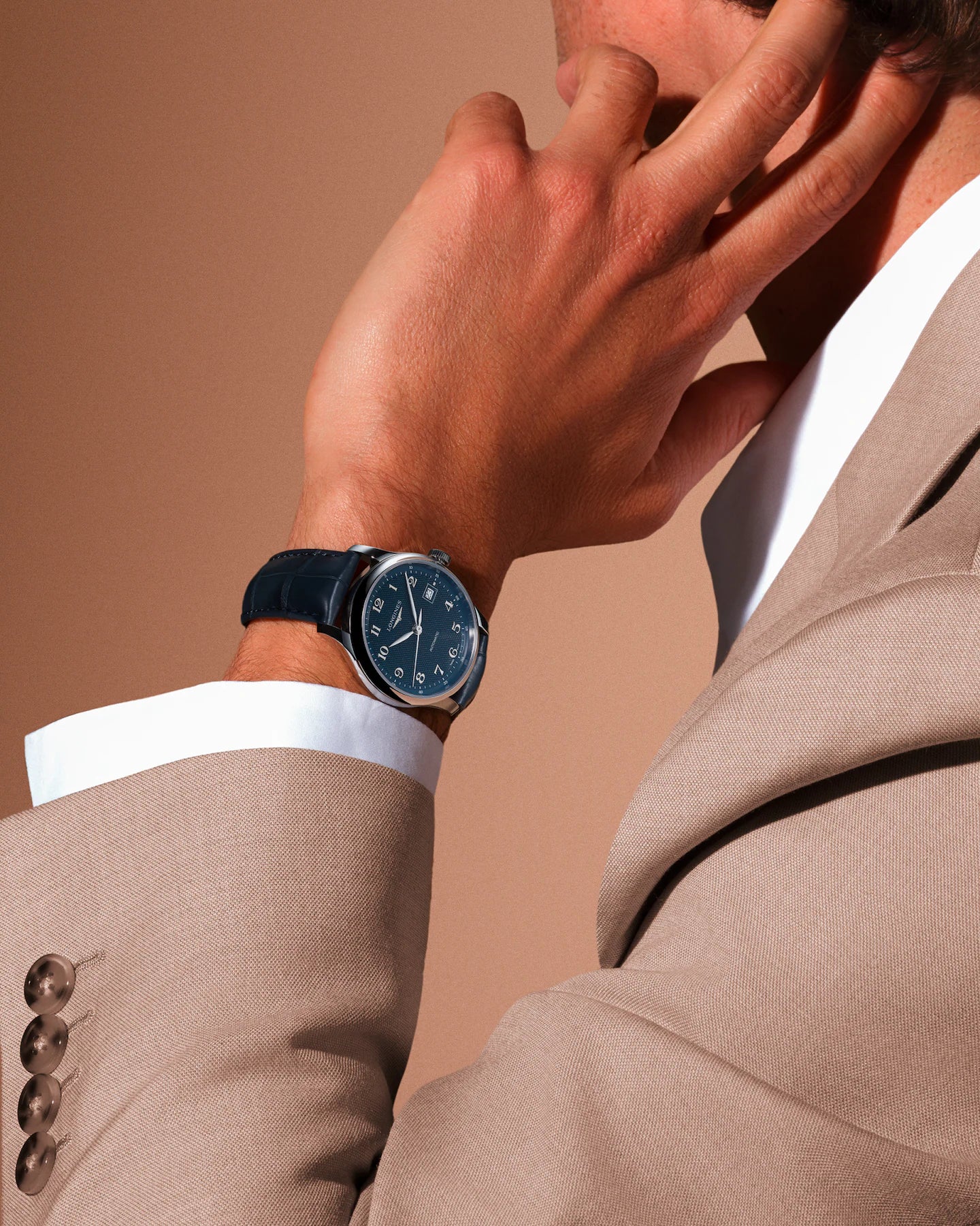 Person wearing a watch with a beige suit and white shirt on a brown background