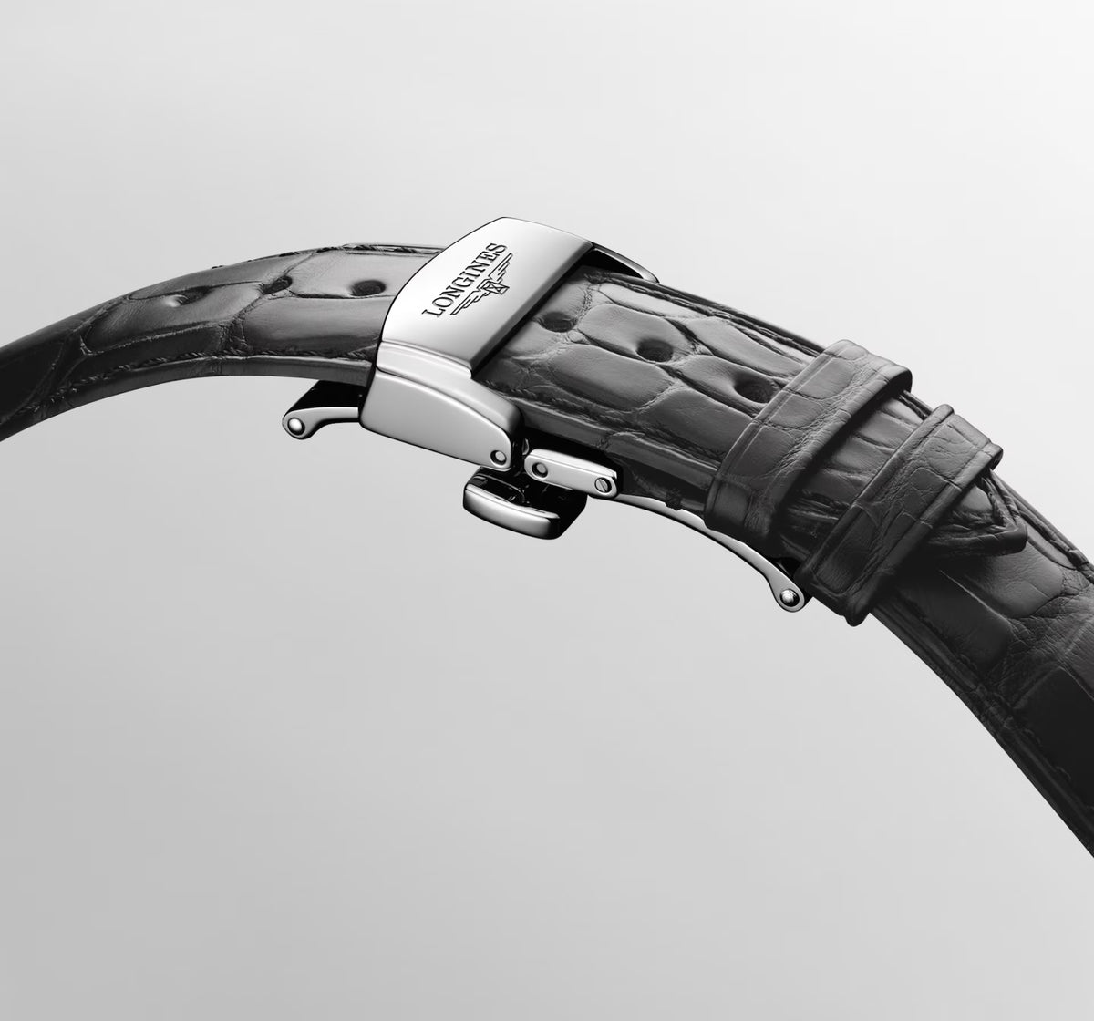 Close-up of a black alligator leather watch strap with a polished silver-toned metal buckle engraved with the Longines logo, set against a light gray background.