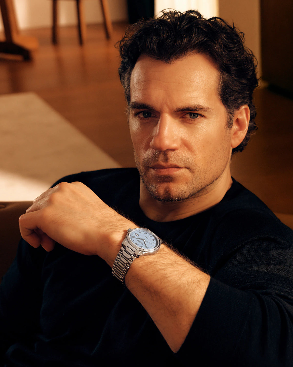 A man wearing a silver-toned metal link watch with a light blue dial, posed in a warm indoor setting.