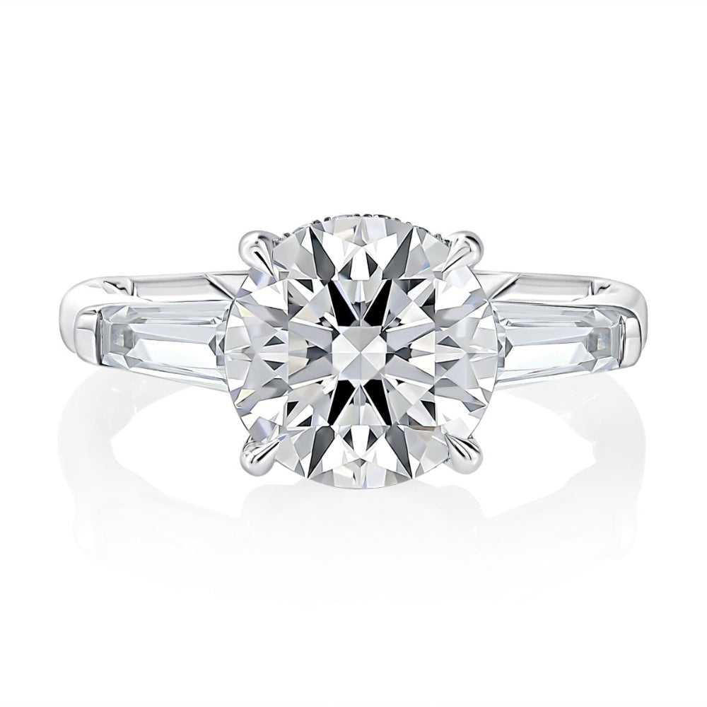A classic round-cut diamond solitaire engagement ring with baguette side stones in a silver-colored metal setting, shown on a clean white background with a soft reflection.