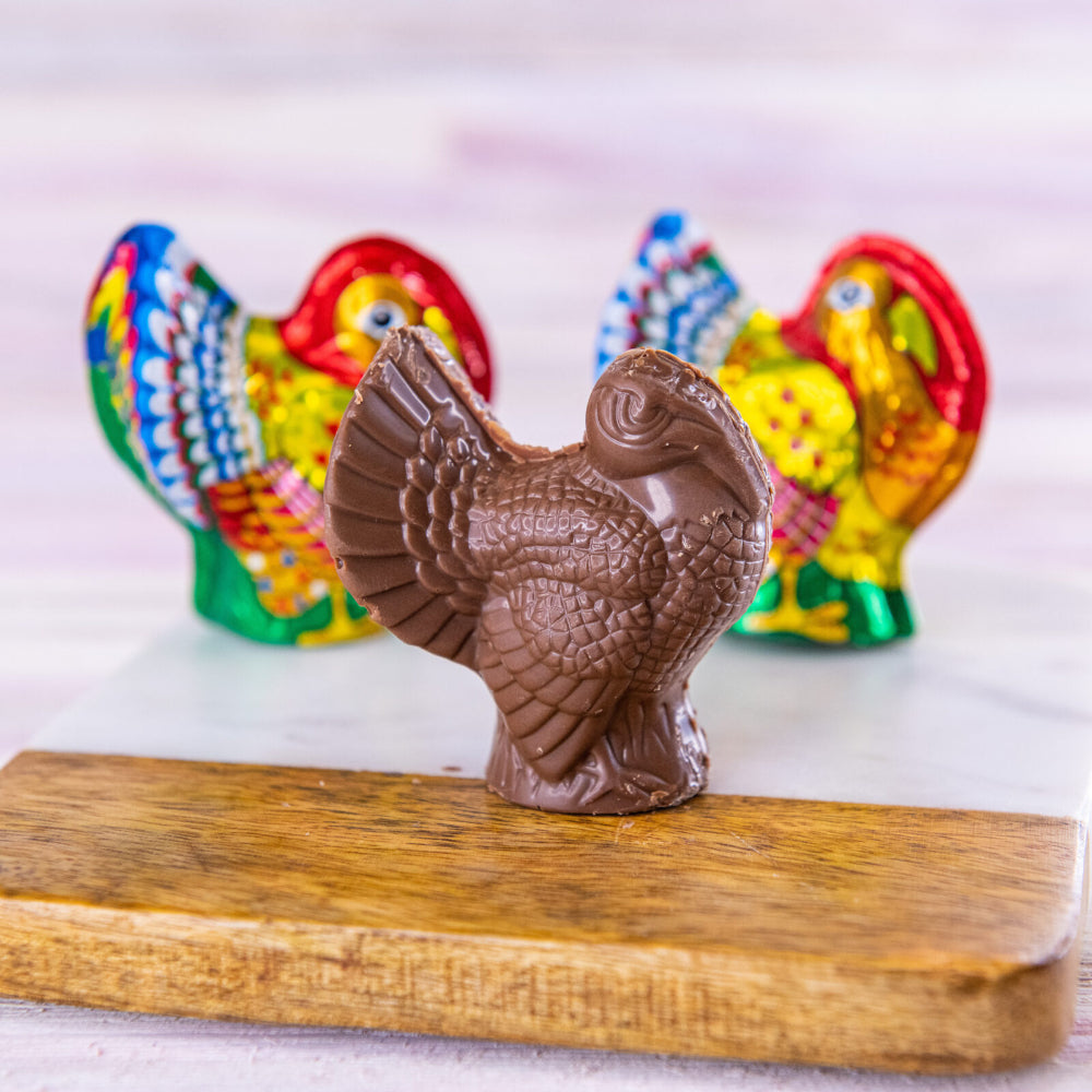 A molded chocolate turkey standing upright on a wooden board, with two colorful decorative turkeys in the background on a light surface.