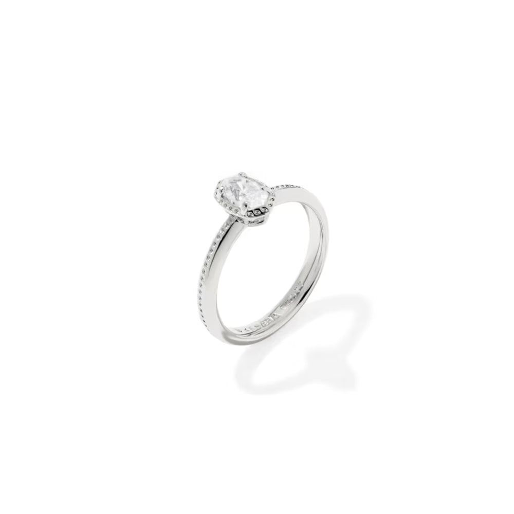 A silver engagement ring featuring a round-cut clear gemstone in a prong setting, with a delicate milgrain pattern on the band, shown on a clean white background with a soft shadow.