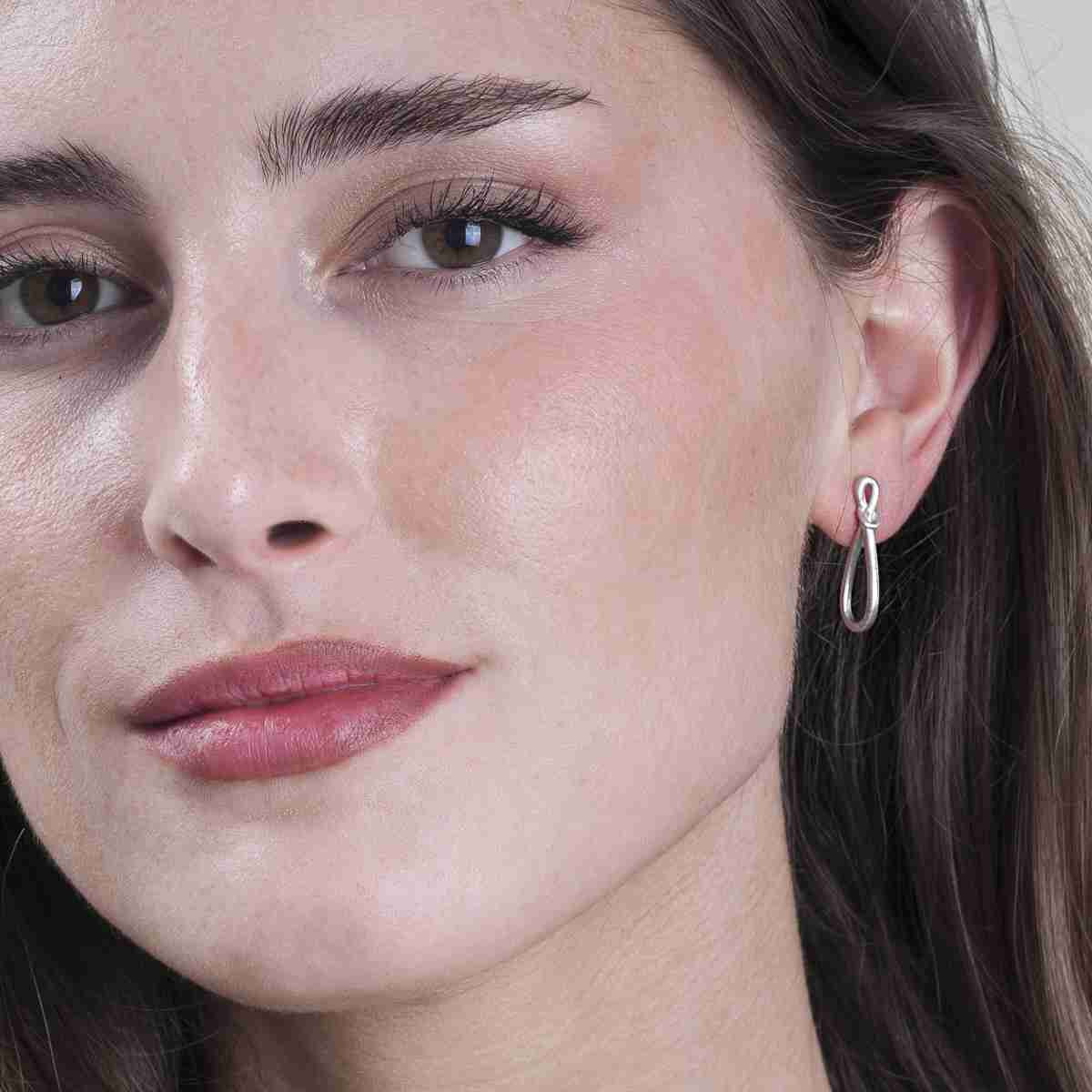 A close-up of a woman's face wearing a delicate silver drop hoop earring with a small curved accent at the top.