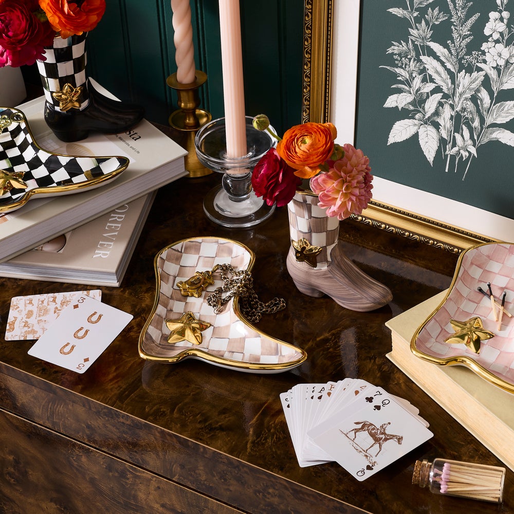 A styled tabletop scene featuring decorative cowboy boot-shaped dishes with a pink-and-white checkered pattern and gold trim, holding cards and jewelry. The setting includes flowers, candles, books, and framed botanical wall art in an elegant interior.