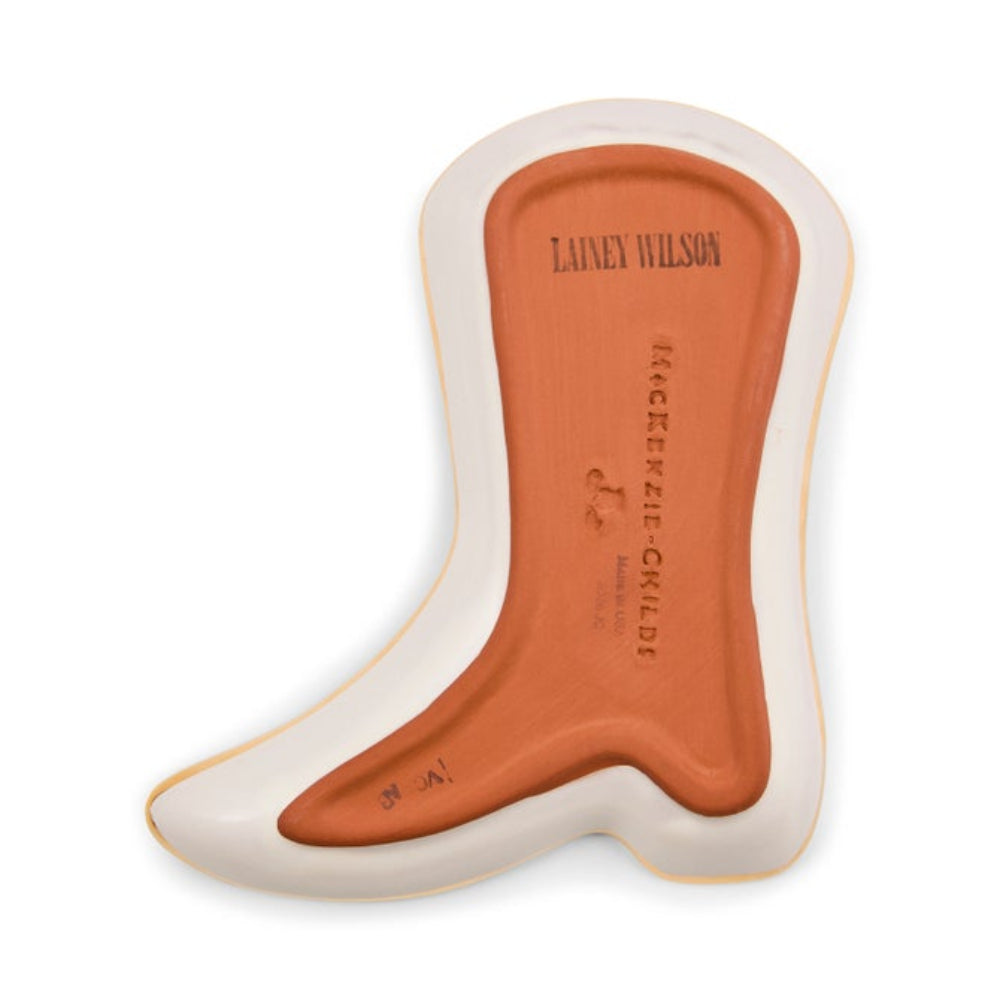 A single boot-shaped product shown on a plain white background, with a warm orange center and a cream outer border. The center is embossed with the text 'LAINEY WILSON' and 'MACKENZIE CHILDS'.
