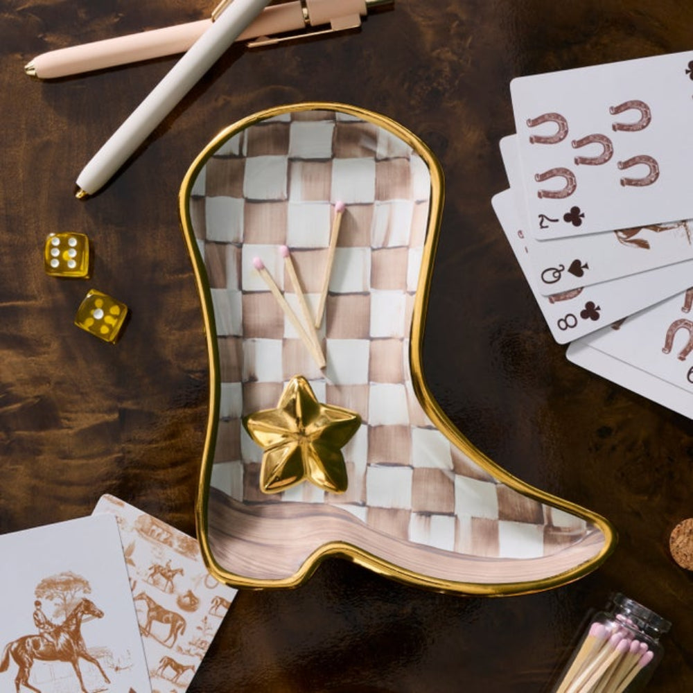 A cowboy boot-shaped matchstick holder with a woven checkered pattern in beige and white, a gold rim, and a gold star topper, styled on a dark wooden tabletop with cards, dice, and matchsticks.