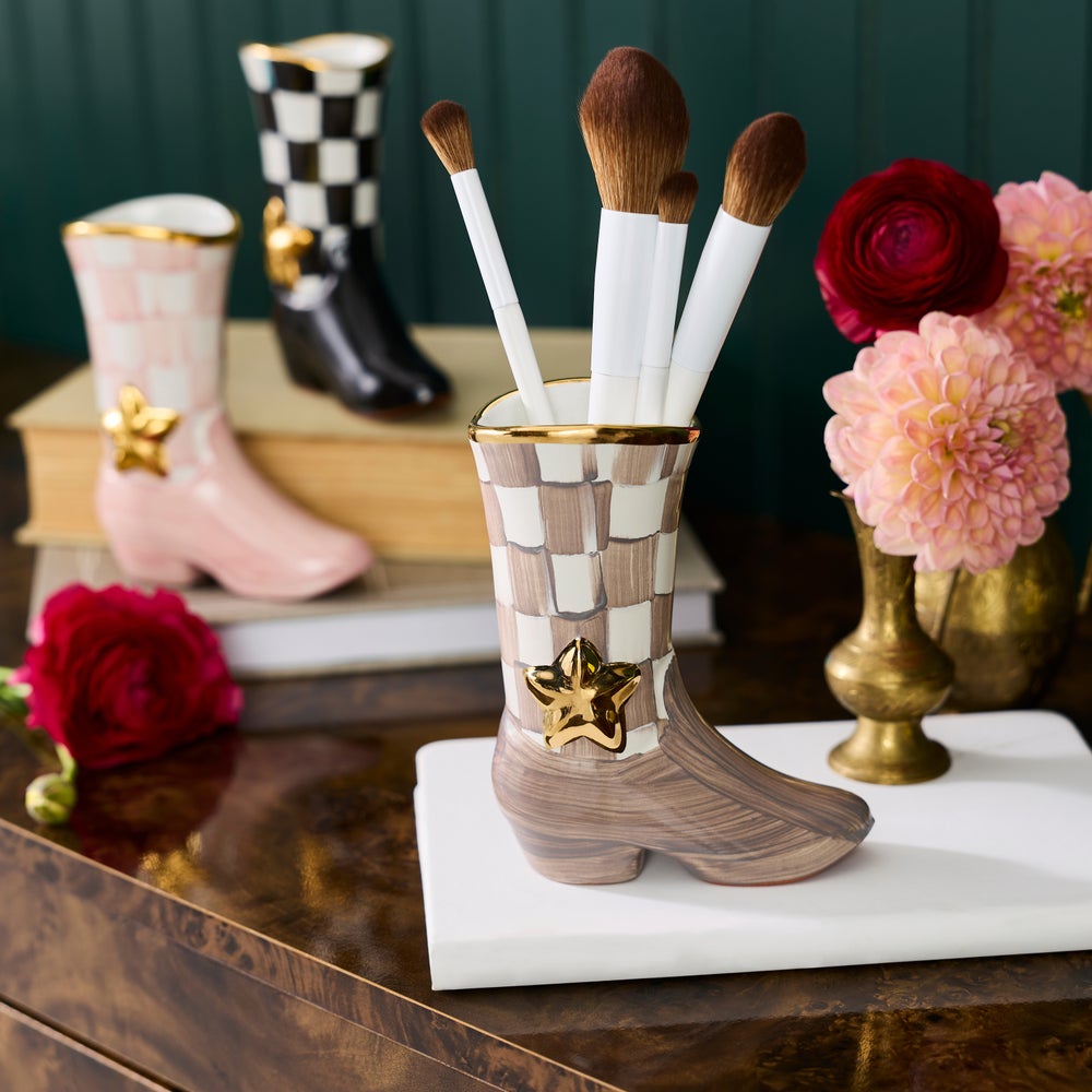 A ceramic vase shaped like a western boot, filled with white-handled makeup brushes, displayed on a white marble tray on a dark wooden table. The boot has a brown and cream checkerboard pattern with a gold star accent.