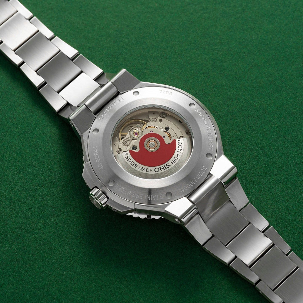 Close-up of the back of a stainless steel wristwatch with a transparent case showing the Oris Big Sea-Mean mechanical movement, set against a textured green background.