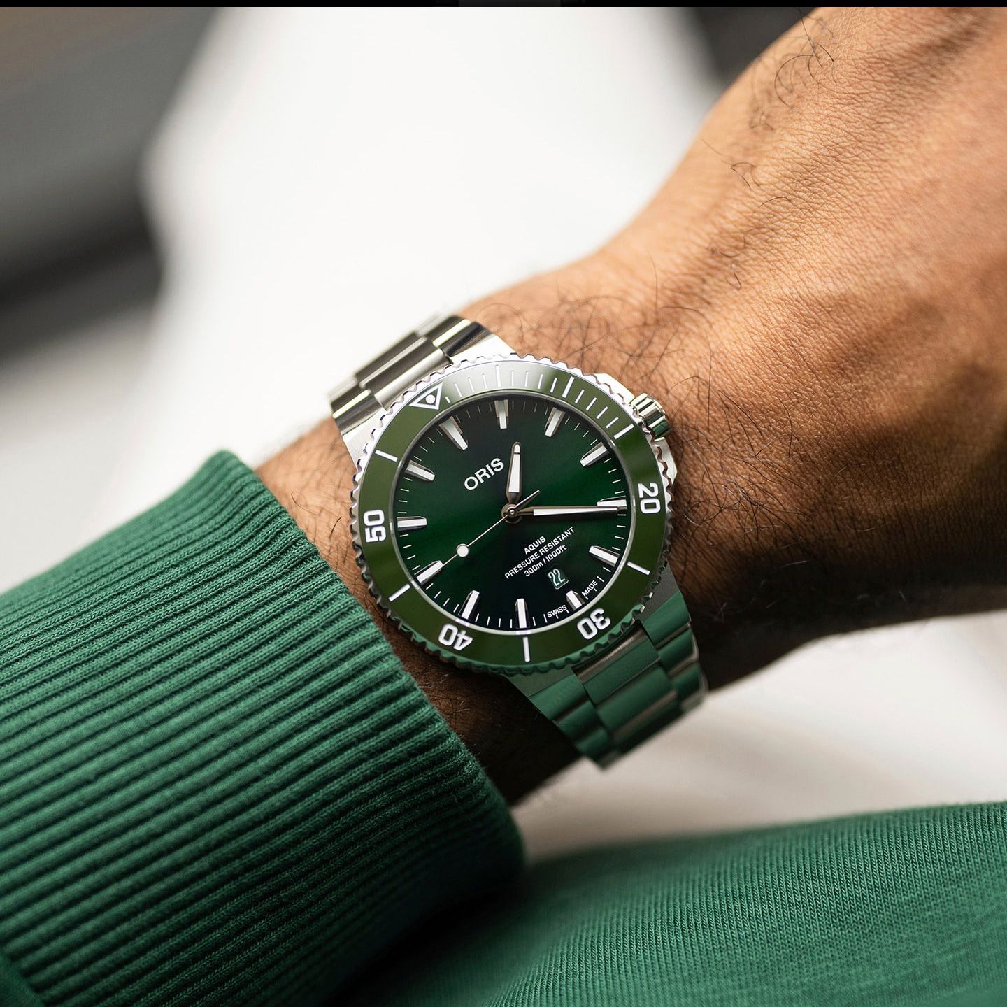 A wrist wearing an Oris Submariner Date watch with a green dial, green bezel, and green metal bracelet, paired with a green ribbed knit sweater sleeve.