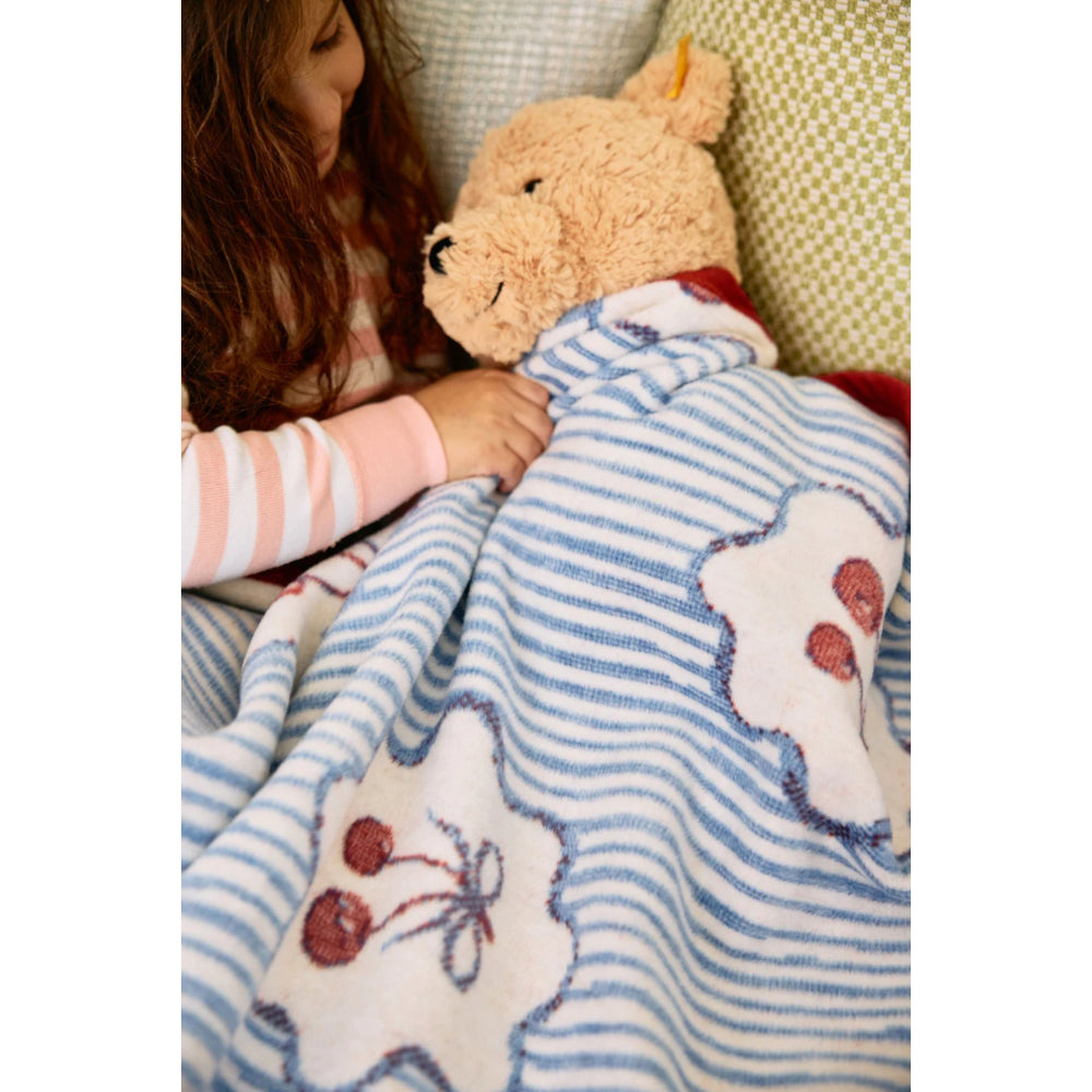 Child holding a teddy bear under a blue and white striped blanket with floral and berry patterns.