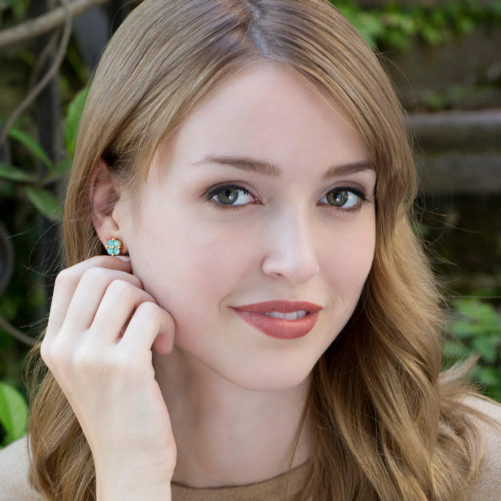 A woman with light brown hair wearing a small turquoise and gold stud earring, touching her ear with her hand. She is wearing a beige top against a soft-focus outdoor background of green foliage.
