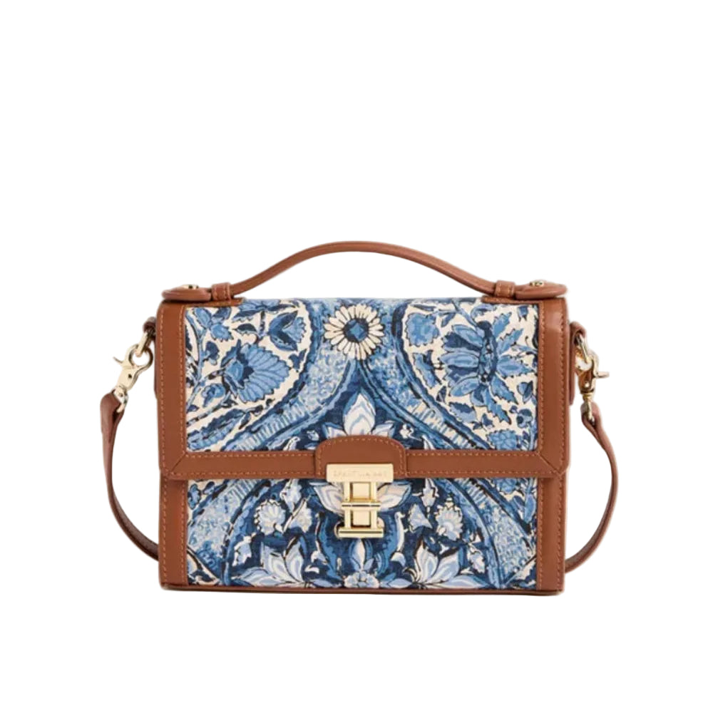 A structured handbag with a brown leather trim and handle, featuring a blue and white floral patterned front panel and a gold-tone turn-lock clasp, set against a solid cream background.