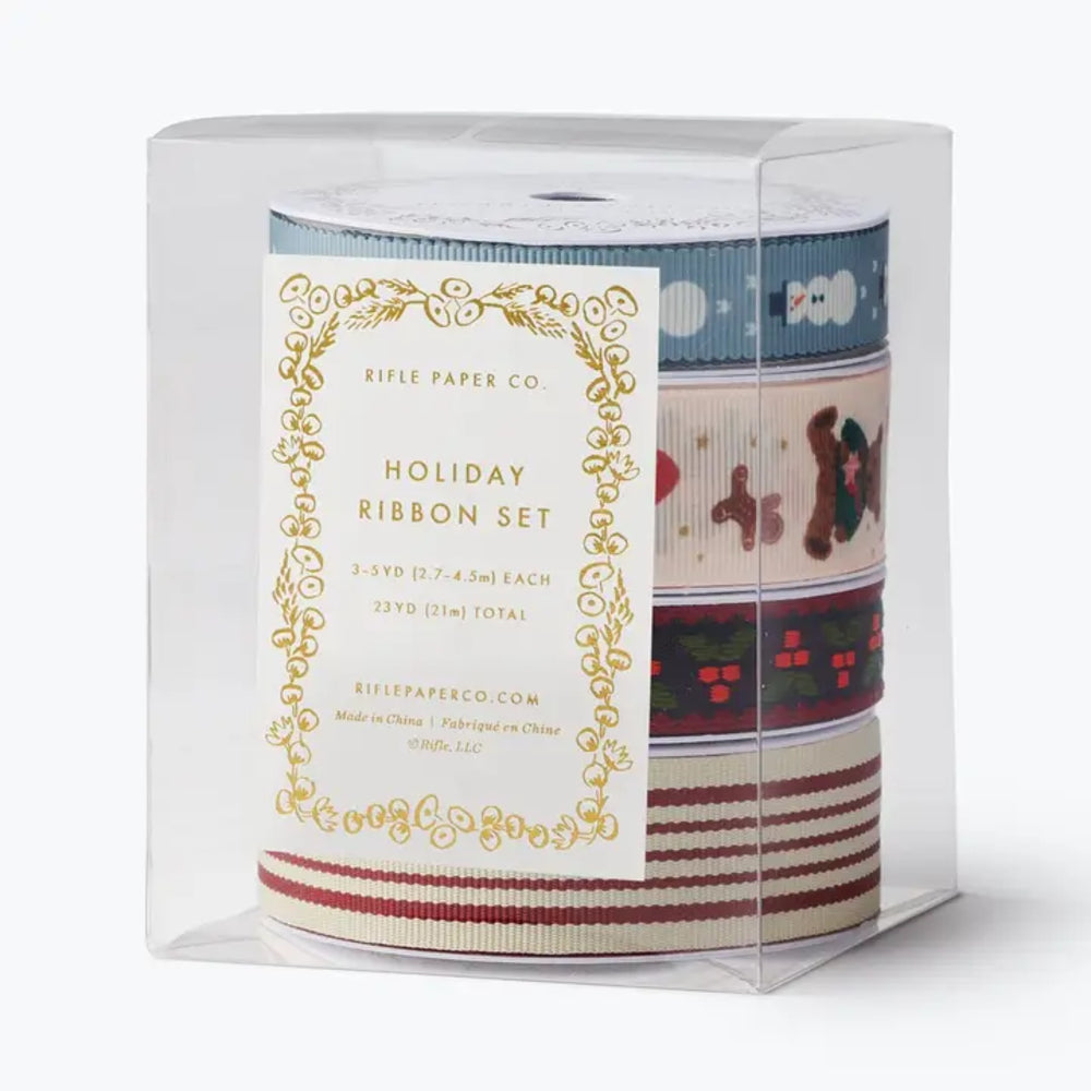 A clear plastic cube package containing a holiday ribbon set with three rolls of patterned ribbon visible behind a white paper label. The ribbons show Christmas motifs including snowflakes, gingerbread cookies, and red accents.