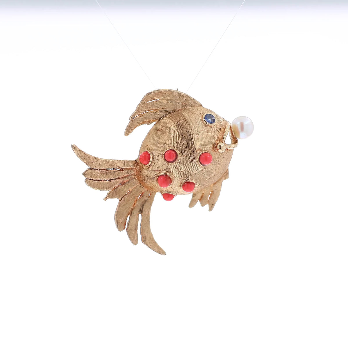 A gold fish-shaped brooch featuring coral stones on its body, a small blue sapphire for an eye, and a white pearl protruding from its mouth, set against a plain white background.