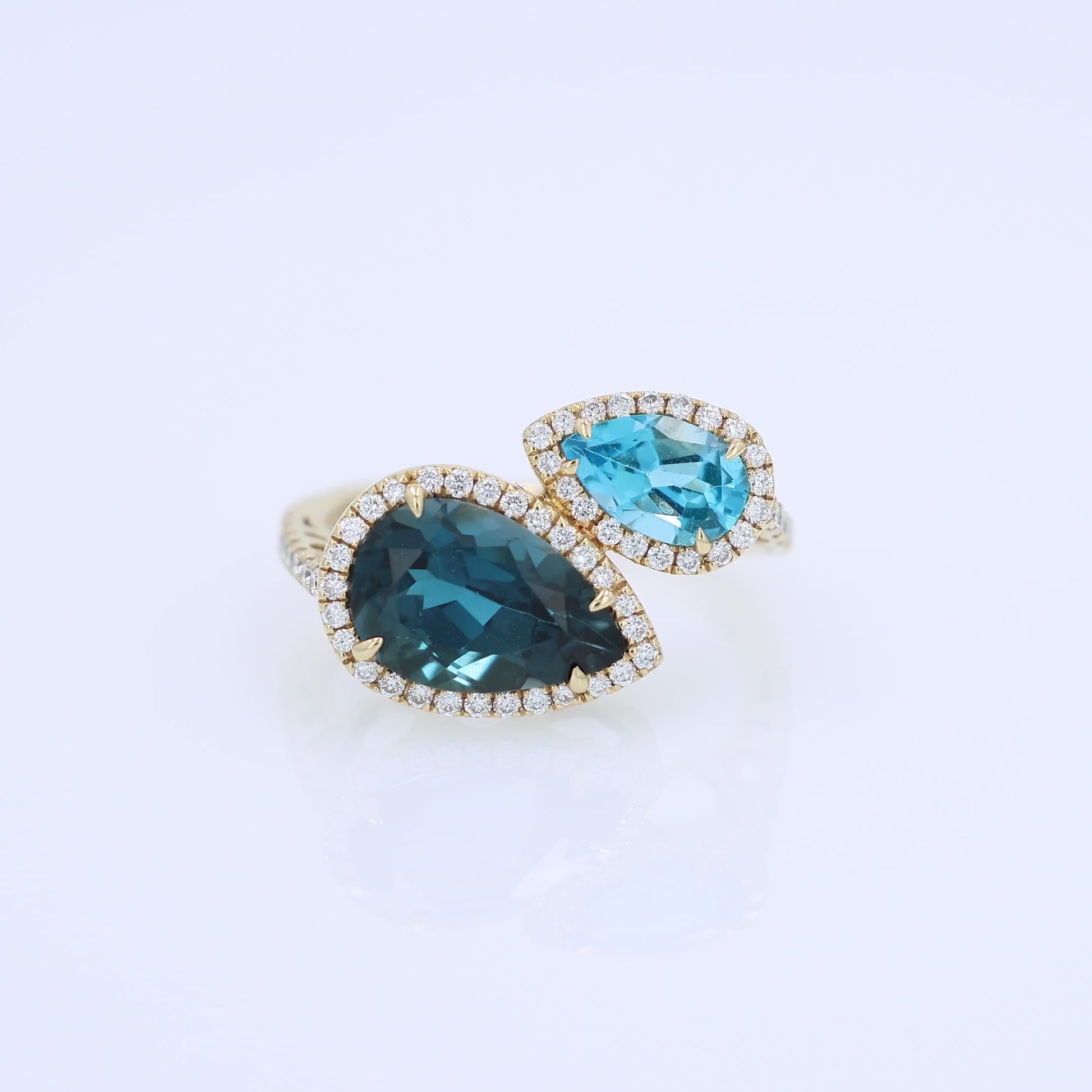 Yellow gold diamond halo bypass ring with 2 pear shape blue topaz on a white background