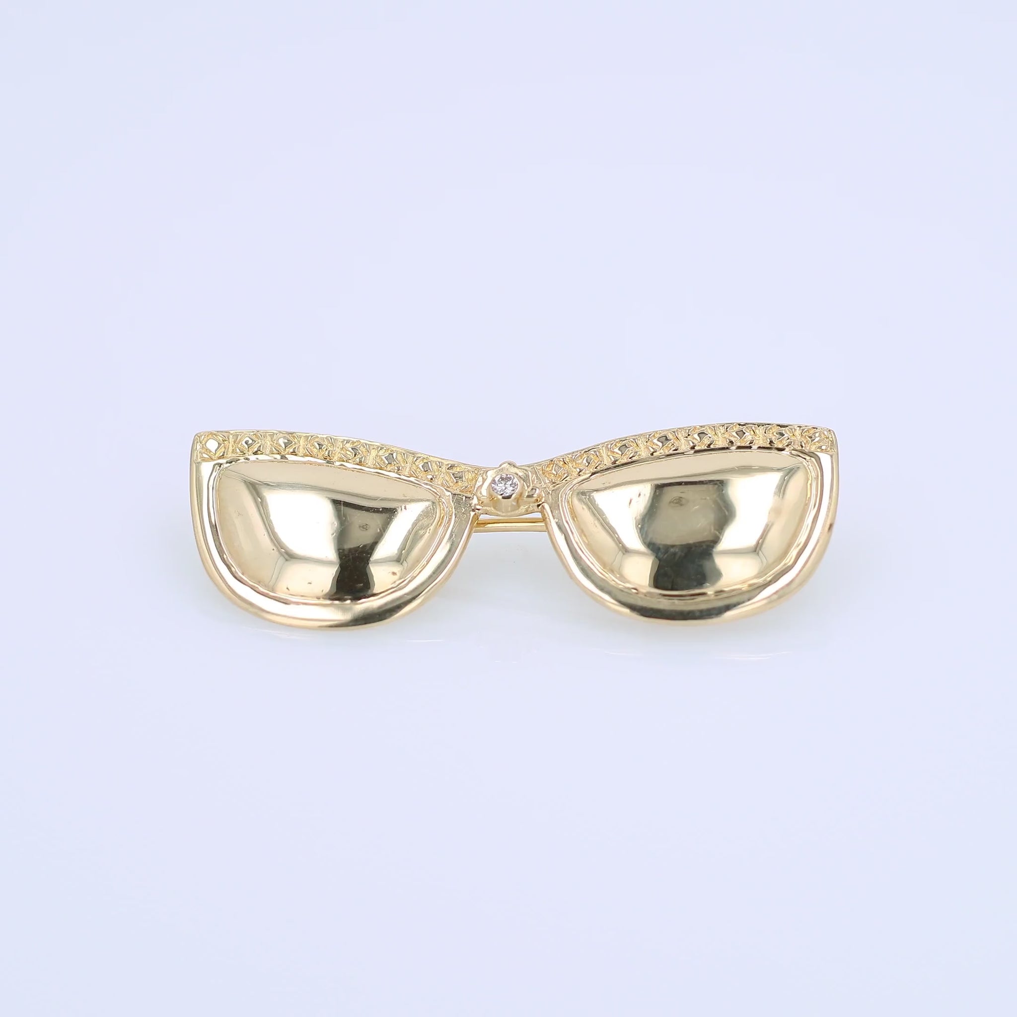 A gold brooch depicting sunglasses with a row of textured gold along the top frame and a central diamond, shown on a plain white background.