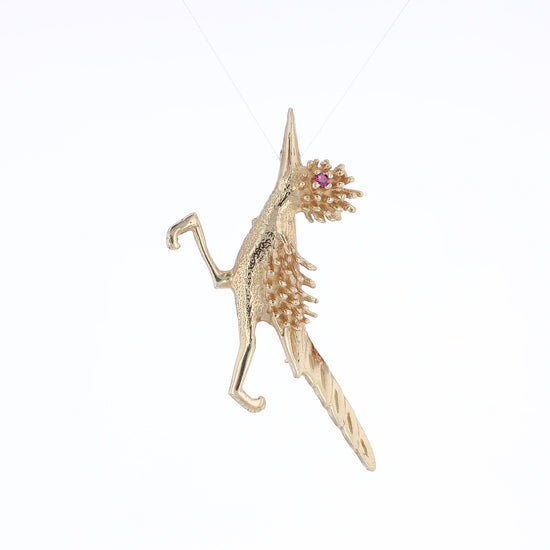 Yellow gold road runner pin with a round pink sapphire eye on a white background