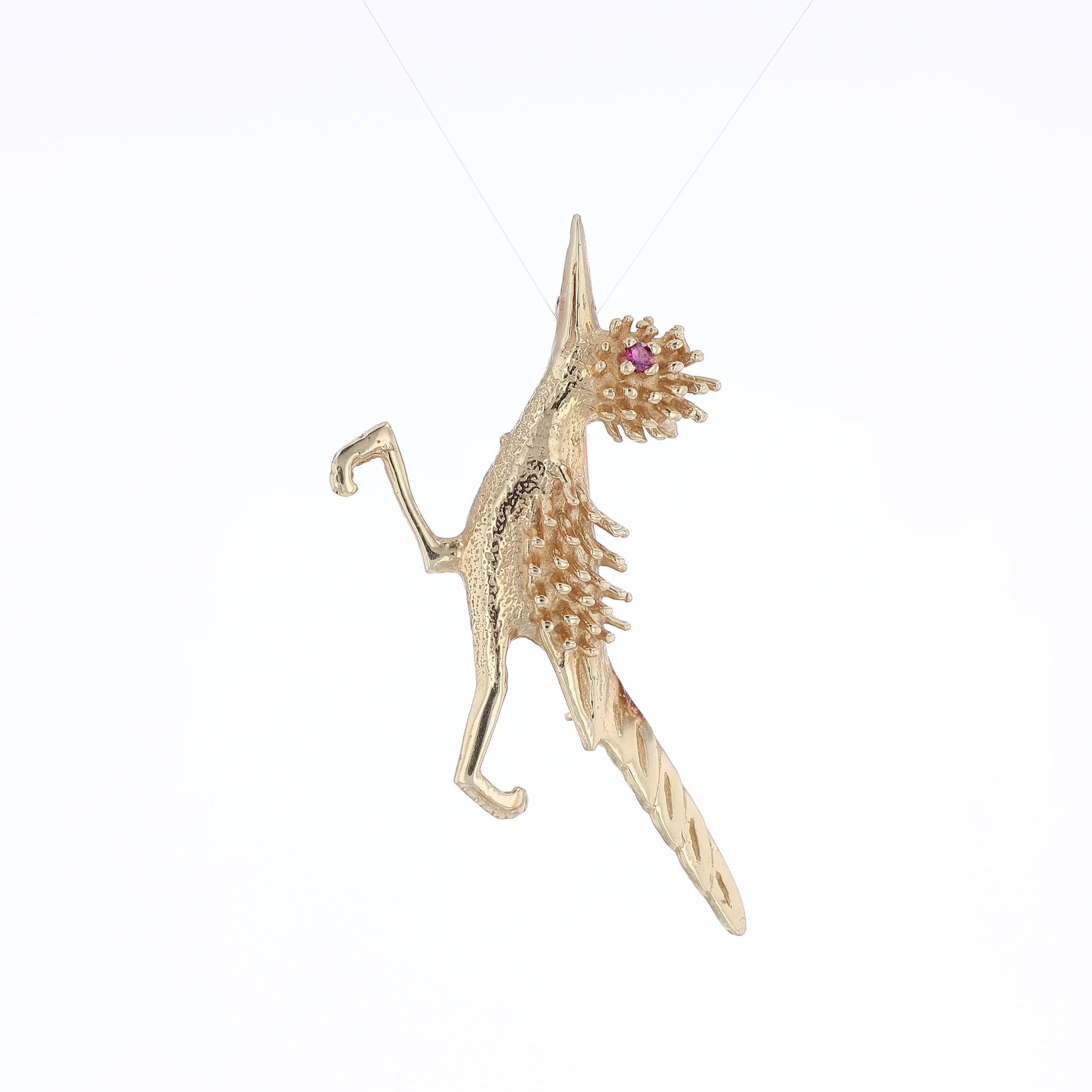 Yellow gold road runner pin with a round pink sapphire eye on a white background