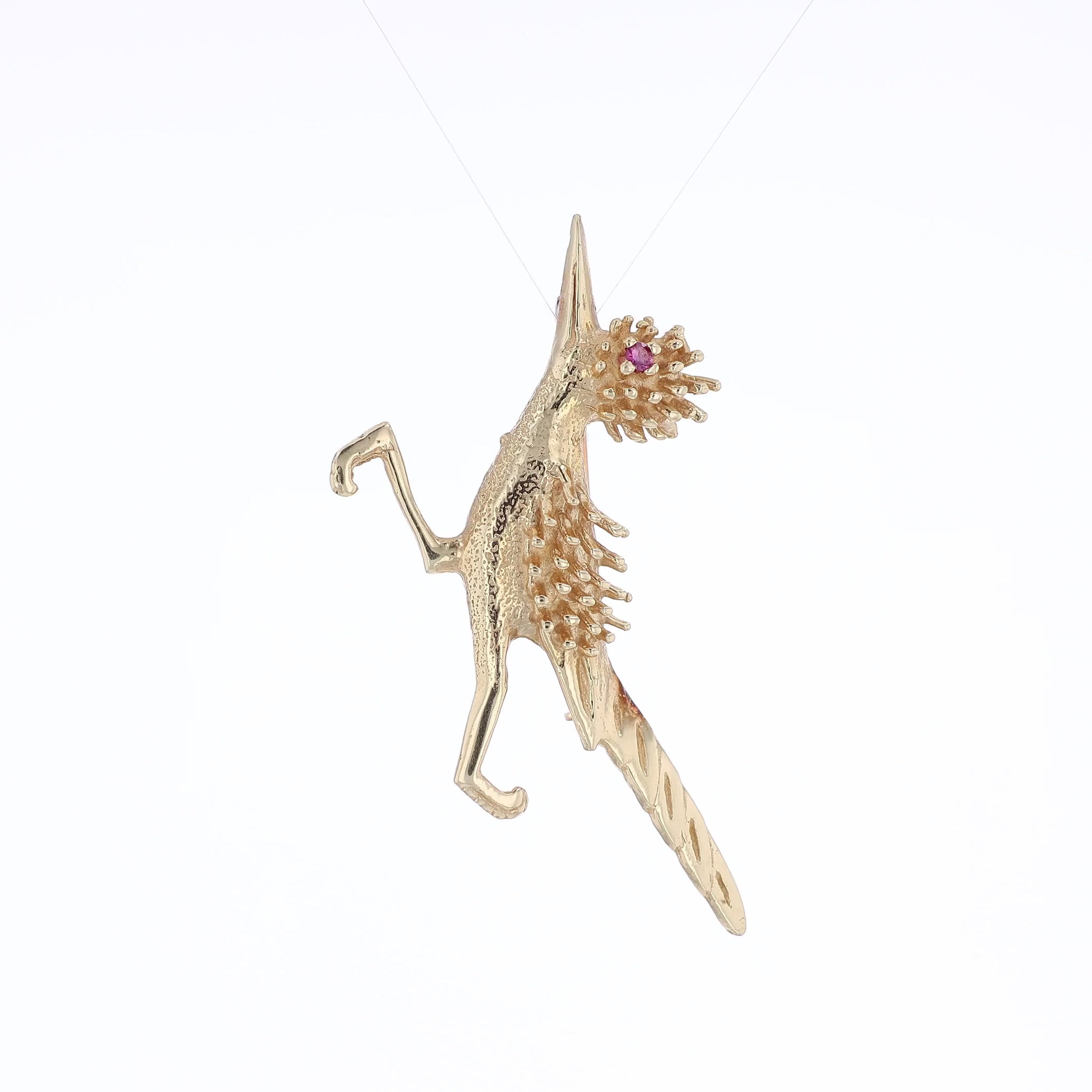 Yellow gold road runner pin with a round pink sapphire eye on a white background