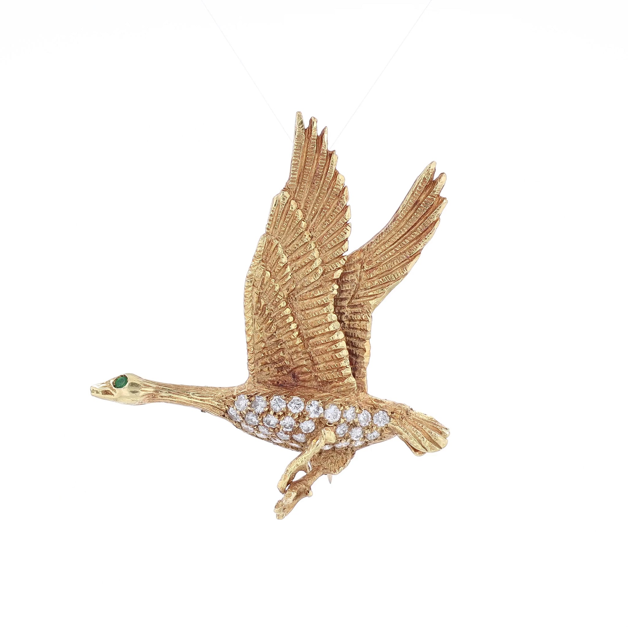 Brushed yellow gold pin of a flying duck with an emerald eye and diamond body on a white background