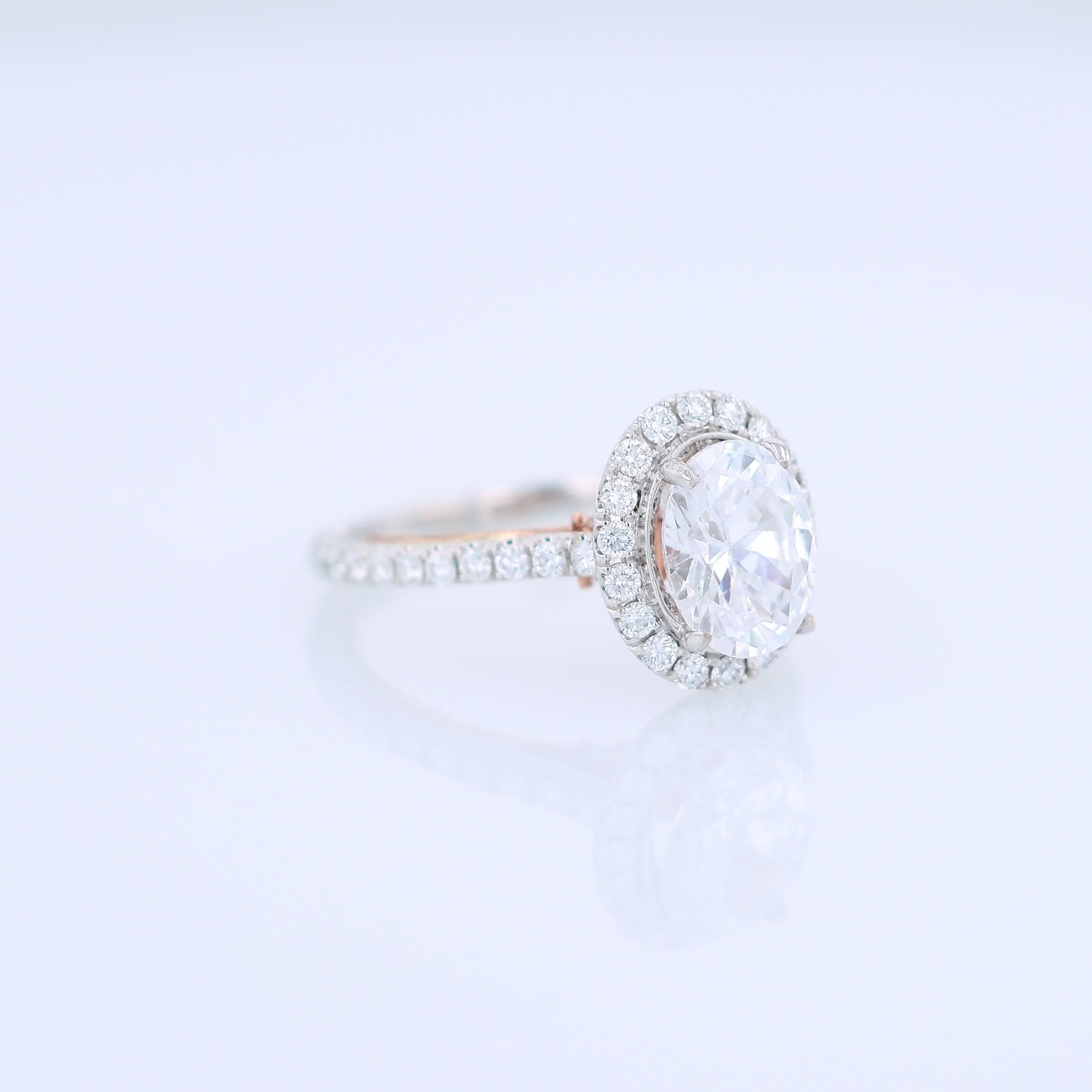 White and rose gold and diamond halo engagement ring with diamond accents and an oval cubic zirconia center on a white background
