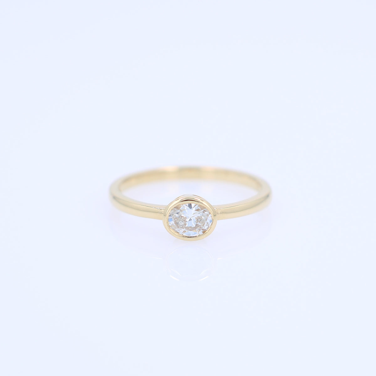 A delicate yellow gold ring featuring a bezel-set oval diamond, shown against a plain white background.