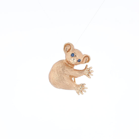 Yellow gold brushed koala pin with two round blue sapphire eyes on a white background