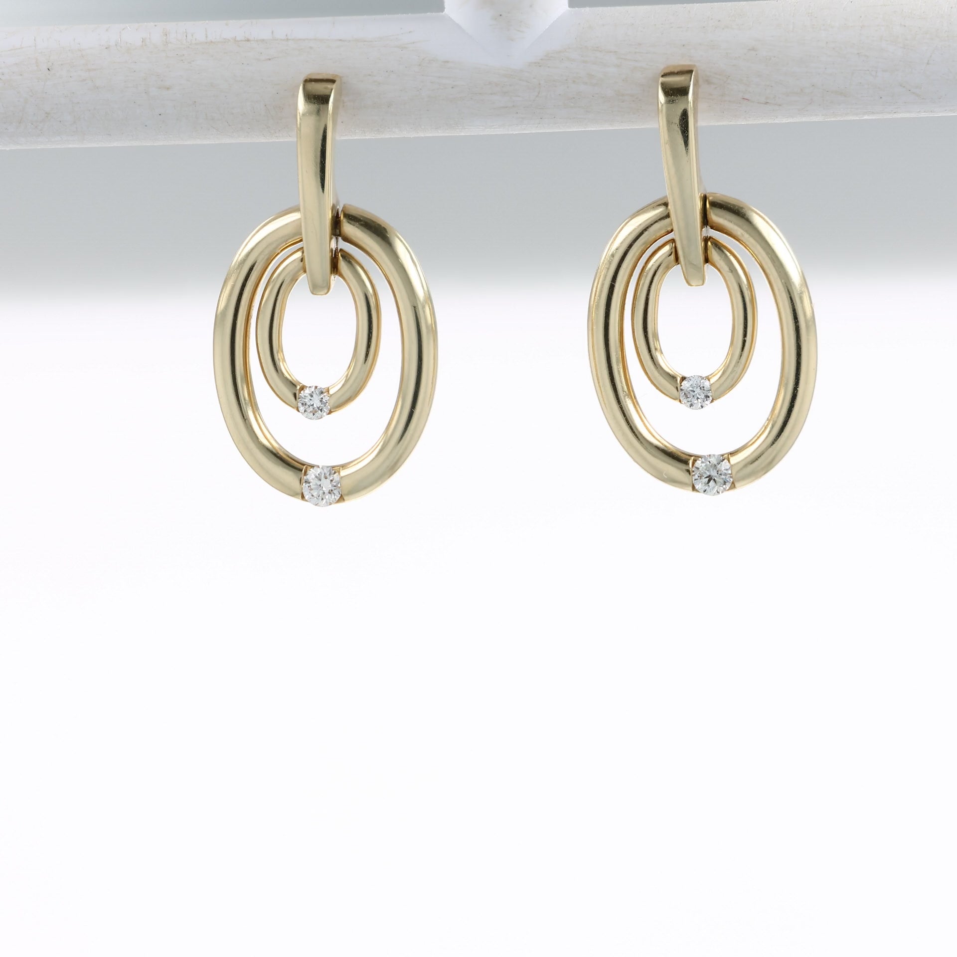 A video of A pair of gold drop earrings with interlocking oval rings and small clear gemstone accents, shown at various angles suspended from an off-white bar on a white background