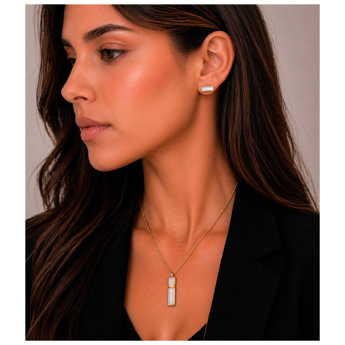 A woman in a black blazer is shown in profile wearing a delicate gold necklace with a vertical rectangular white stone pendant and a matching small rectangular stud earring.