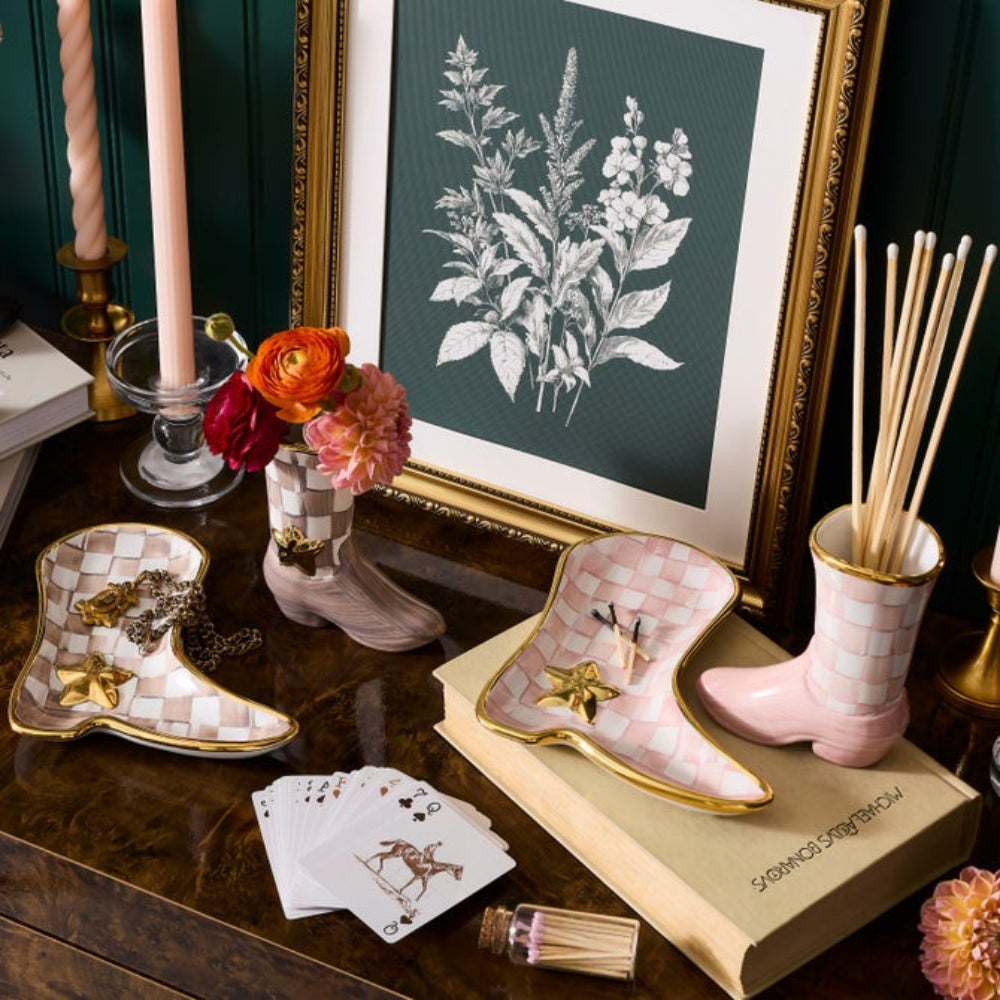 A styled tabletop scene featuring two boot-shaped candle holders with pink-and-white checkered patterns and gold trim, one holding a candle and the other a reed diffuser. A matching boot-shaped dish sits beside them.