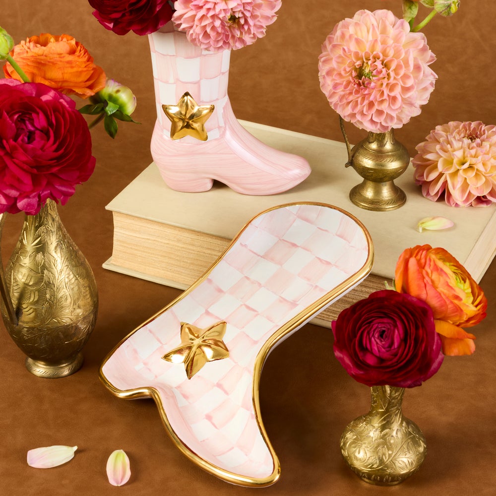 A decorative boot-shaped dish or tray in pale pink with a quilted pattern and gold trim, styled on a brown surface with flowers and small vases around it.