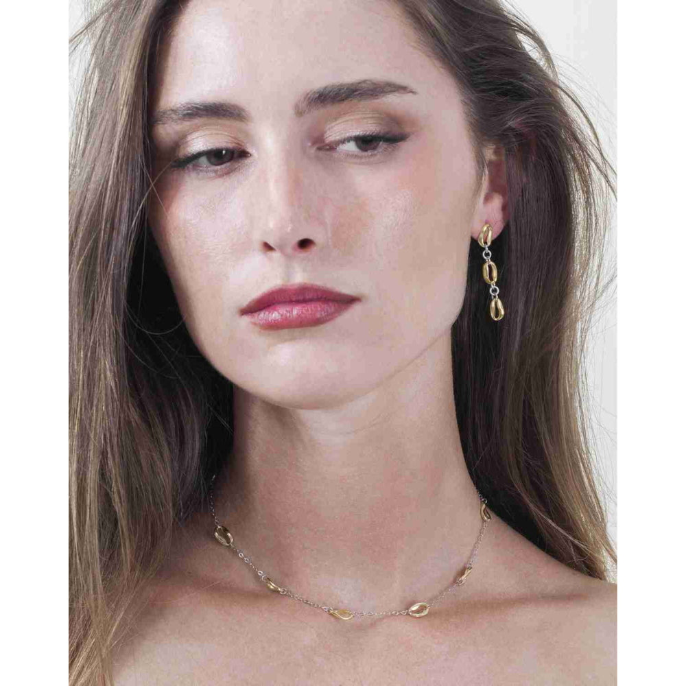 A close-up portrait of a woman wearing delicate gold jewelry, featuring a thin gold chain necklace and a matching gold drop earring with a small gold bead.