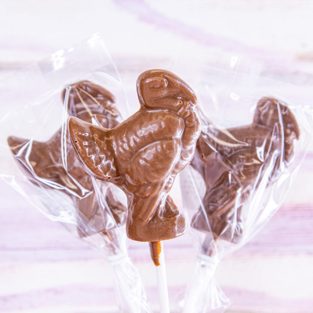 Three chocolate lollipops shaped like turkeys individually wrapped in clear cellophane packaging on white sticks, set against a soft pink and white background.