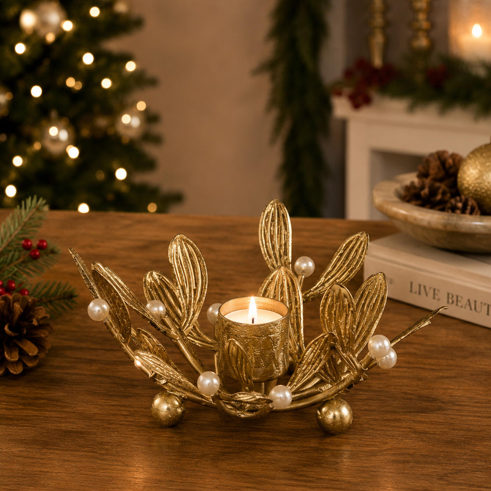A gold-toned leaf-shaped candle holder with pearl accents and small metallic gold balls, holding a lit tealight candle on a wooden table. A blurred Christmas tree and holiday decor appear in the background.