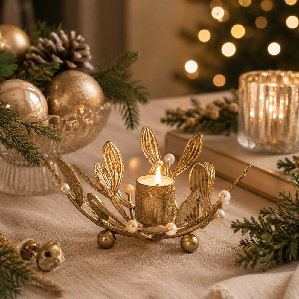 A gold-toned leaf-shaped candle holder with small pearl-like beads and metallic gold finials, holding a lit tealight candle on a table. The scene is decorated with pine branches, gold ornaments, a glass bowl, and a blurred Christmas tree in the background.