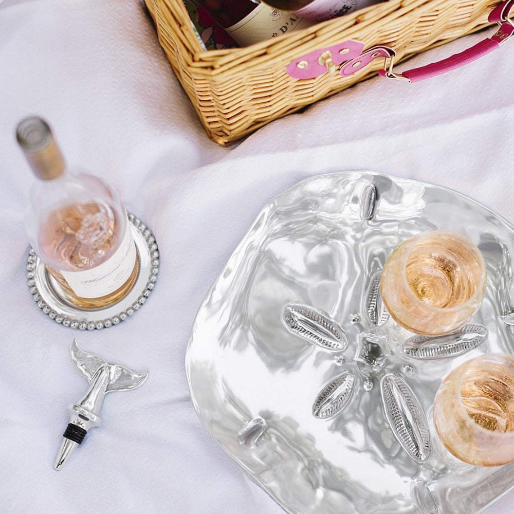 Silver aluminium decorative wine coaster with beaded edge on a white table with a clear wine bottle on it, with a silver sand dallor platter and silver winged wine bottle topper with refreashments on it and in a brown wicker wine holder chest . 