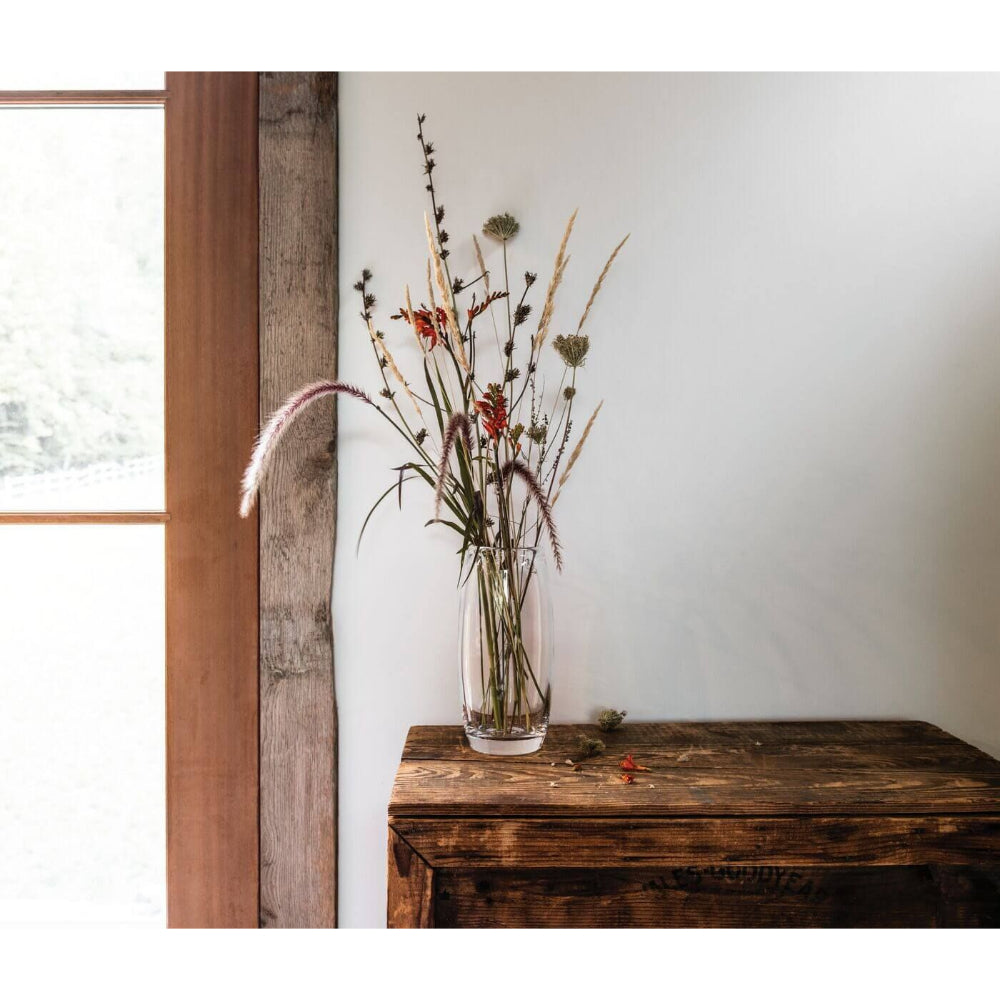 A clear glass vase with flowers in it on a wood table next to a brown pained window.
