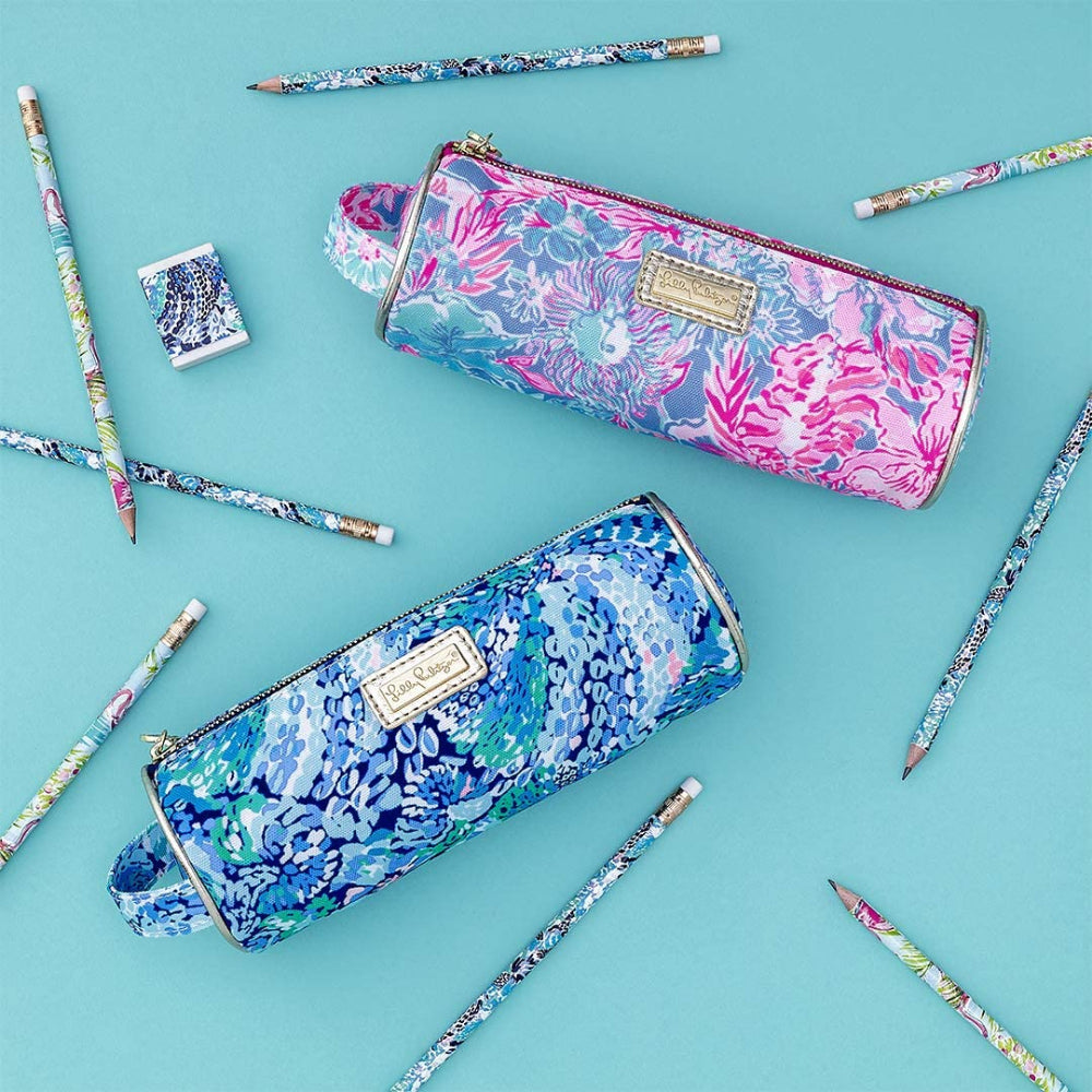 A anged side photo of a  Lilly Pulitzer pencil pouch with a multicolored floral pattern and gold pu accents, featuring a zipper closure and a gold Lilly logo charm zipper pull next to another one in blue with pencils scathered around on a blue backround.