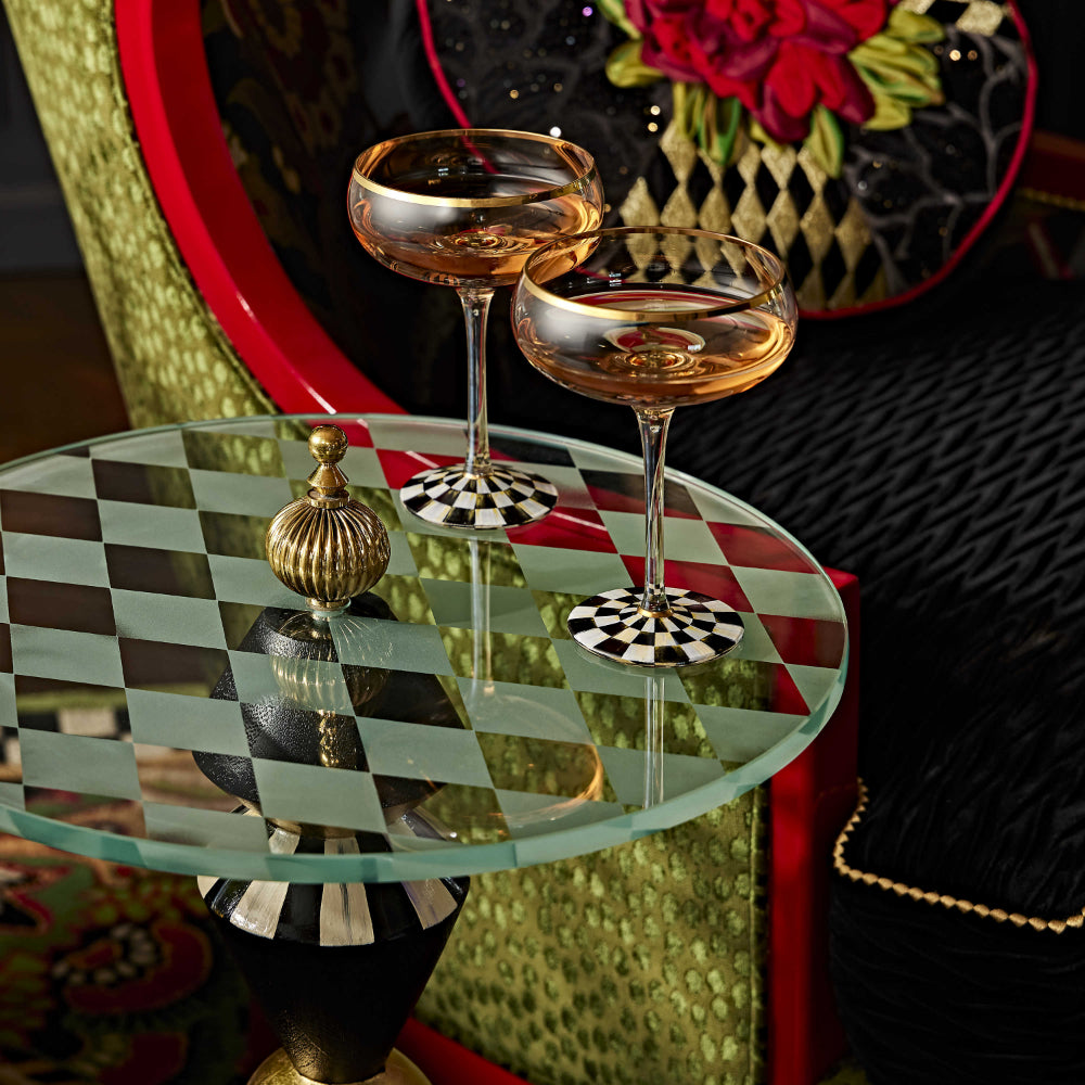 Life style photo of MacKenzie-Childs Gambit Accent Table with wine glasses on table with black painted wall with green chair