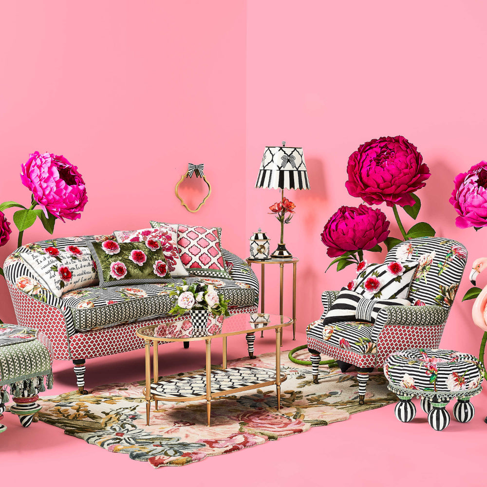 A photo life style photo of a gold-framed glass side table with a decorative white and black lower with flowers and other black and pink furniture on in a pink painted room with a black and white lamp and clock on it.
