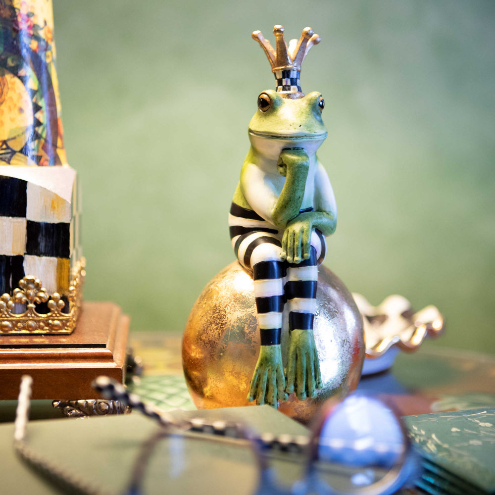 A hand-painted green frog with black and white stripes, sitting on a gold-leafed ball, wearing a crown.