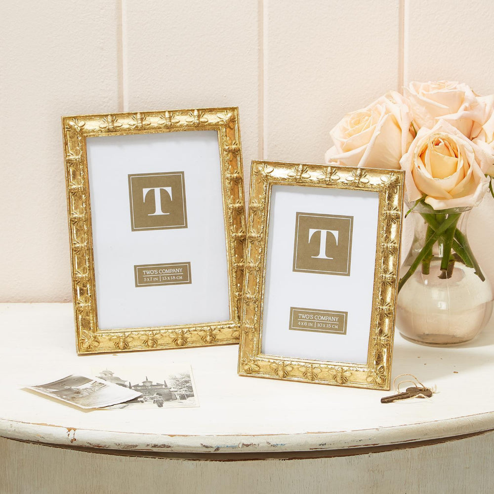 Two gold leaf Napoleon Bee photo frames of different sizes 4x6 & 5x7, capable of standing vertically or horizontally on a white table with a vase with pink flowers in it and a key and photos against a white wall. 