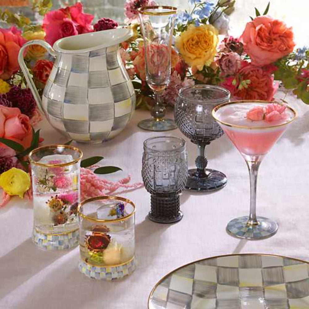 Clear glass highball with gold rim and sterling check patterned base on a white table with other MacKenzie-Childs sterling check pitcher and plate and colorful flowers with other drinkware items 