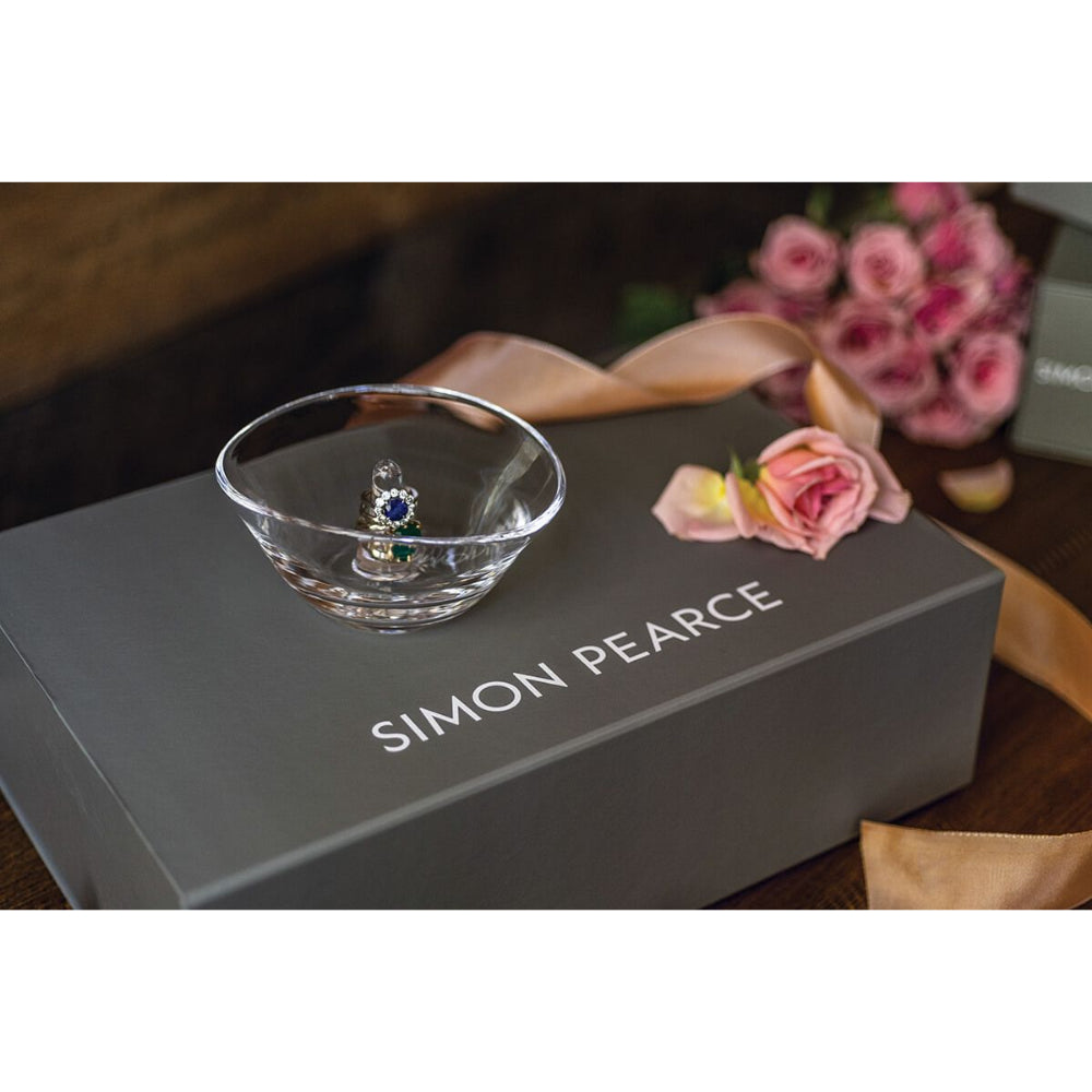 Clear glass ring holder with a unique design on a gray Simon Pearce box on a brown table with a pink rose.