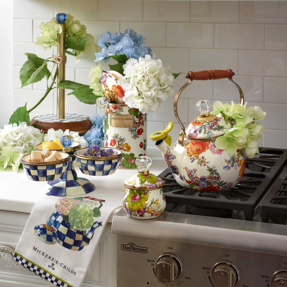 White towel with a royal check floral teapot design on a kitchen counter next to a stove with flowers,tea pot and other royal check items.