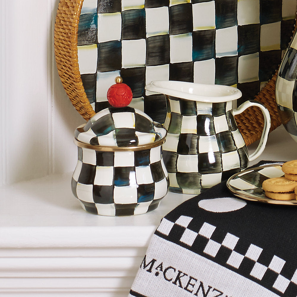 A lidded sugar bowl with a Courtly Check enamel pattern and a red faux-cinnabar bead on the lid handle, rimmed in bronzed stainless steel on a white shelf with other simular items. 