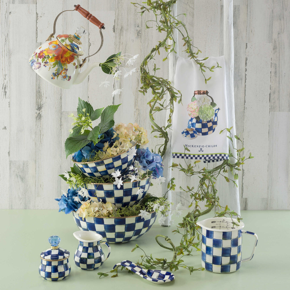 Medium bowl with royal blue and white checkered pattern with other  similar pattered bowels and items with  green vines and other green and blue plants and flowers on a white table with a white curtain backround 