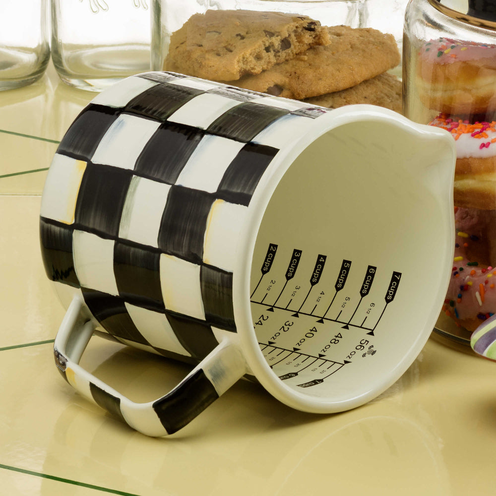 Measuring cup with black and white checkered pattern on a beige colored tile counter to with glass jars with cookies in them. 