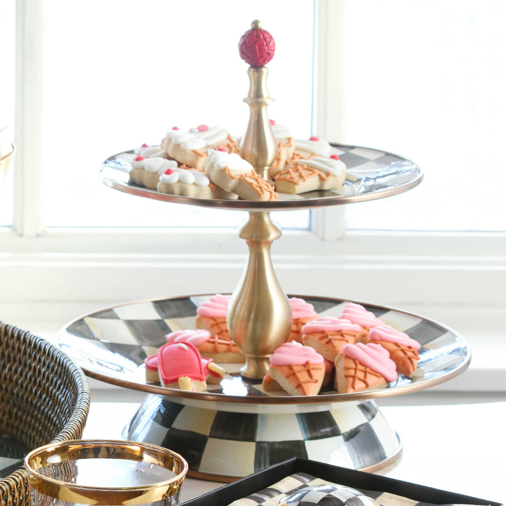 Two-tiered serving tray with black and white checkered design and gold accents on a table with pink and white cockies on it. 